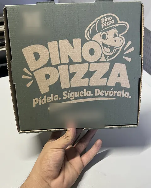 Caja de pizza con fondo verde impreso y un dinosaurio con gorra en la tapa, sostenida en la mano